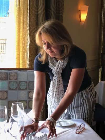 Gia Page, a Caucasian woman with blonde hair and wearing a navy top, navy and white striped pants, and a patterned scarf around her neck, adjusts a table setting next to a sunny window in Davio's Lynnfield
