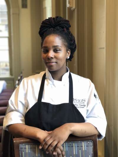 Olisha Sealy, an African American woman with her hair in braids piled atop her head, poses, wearing chefs whites, a black apron, and arms in a relaxed fold over the back of a chair, in the Davio's dining room