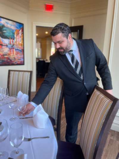 Man, dark hair, beard, dark suit, placing a glass on a table