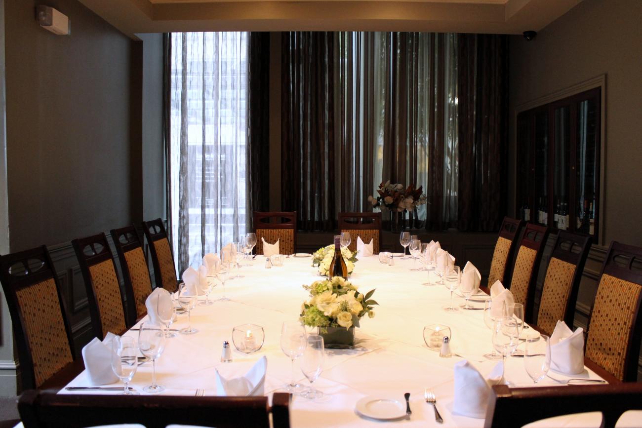 A room with glass-doored wine refrigerators in the walls and sheer curtains for privacy on one end. A long table is set for 16 and surrounded by high-backed chairs with upholstered backs