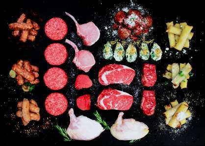 Raw steaks, burgers, pork chops, sausages, and chicken, plus oysters Rockafellar, spring rolls sprinkled with parmesan, and meatballs in marinara, all arranged on a chalkboard background