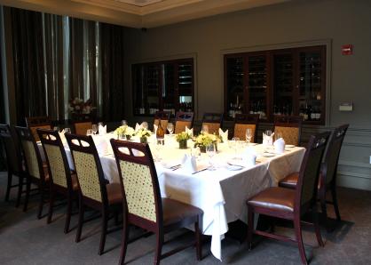 A room with glass-doored wine refrigerators in the walls and sheer curtains for privacy on one end. A long table is set for 16 and surrounded by high-backed chairs with leather seats