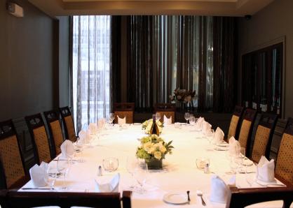 A room with glass-doored wine refrigerators in the walls and sheer curtains for privacy on one end. A long table is set for 16 and surrounded by high-backed chairs with upholstered backs