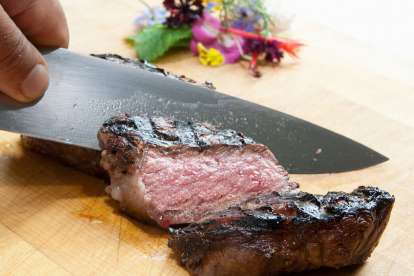 A hand holds a chef's knife carving a medium rare steak