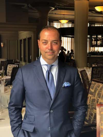 Armando Dias, wearing a gray suit, pale shirt, blue tie, and pocket square, stands with hands in pockets in the Davio's dining room