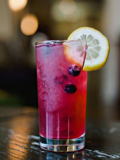 A highball glass with a magenta cocktail garnished with fresh blueberries and a lemon round