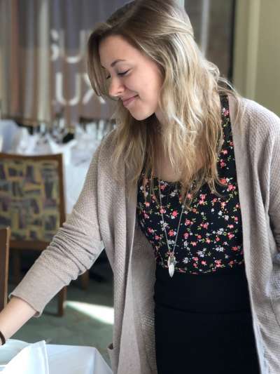 Casey Sarno, a Caucasian woman with long blonde hair and wearing a bloack floral top, black skirt, and beige sweater, adjusts  a table setting in the Davio's dining room 