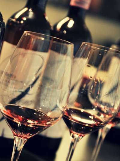 Shown at a slight angle, a row of wine glasses each featuring a few sips of red wine stands in front of a row of bottles of wine. The front left glass is in focus, with the other glasses and bottles becoming increasingly unfocused as they proceed to the right