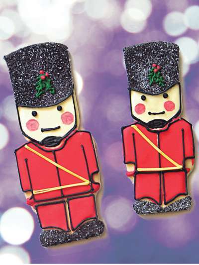 Two sugar cookies decorated as tin soldiers, complete with rosy cheeks and black sugar on their helmets and boots, on an out of focus background of purple and white lights