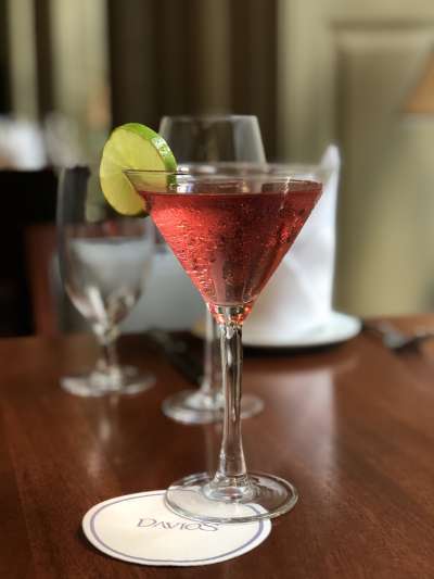 A reddish cocktail in a martini glass with a circular slice of lime on the edge of the glass. The glass sits on a Davio's coaster and the background ofthe restaurant is in soft focus.