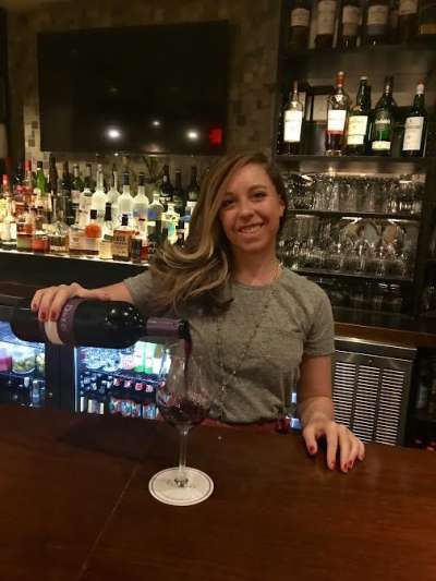 Jennifer Schubert, a Caucasian woman with long hair flowing over her right shoulder, pours wine at the Davio's Manhattan bar while wearing a gray top and long necklace