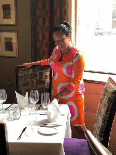 Jess Phifer, wearing a bright orange, white, and hot pink dress, arranges a place setting in the dining room