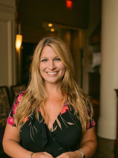 Jessica Newall, a Causasian blonde wearing a v-necked black dress with bright colored flowers on the sleeves, stands smiling in a Davio's dining room