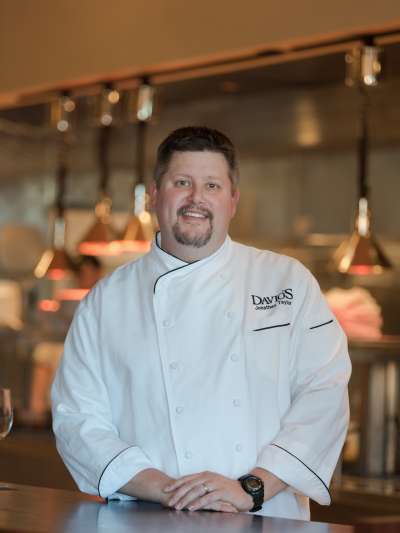 Jonathan Taylor, wearing  chef's whites, poses in the Davio's kitchen. Metal appliances and low-hanging lights are in soft focus in the background.