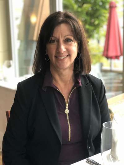 Julie Millard, a Caucasian woman with shoulder-length brown hair, sits wearing a black jacket and a dark purple top with a gold zipper at the neck, the Davio's King of Prussia patio visible in soft focus through the window behind her