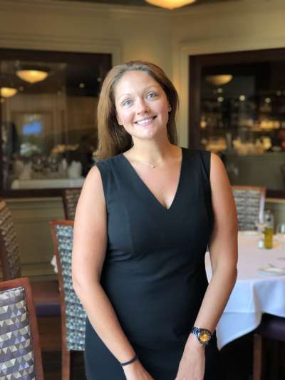 Kara Dunbar stands in the Davio's dining room, smiling and wearing a tailored black v-neck dress