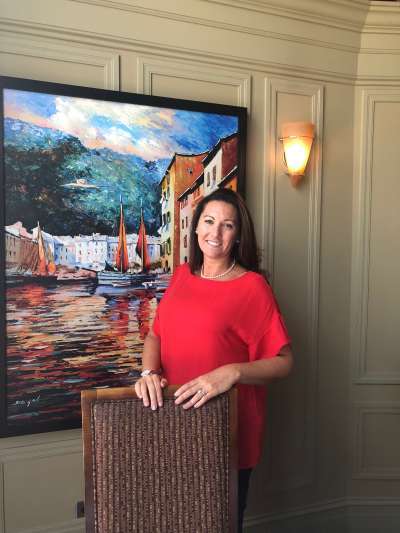 Kate Cunningham, a Caucasian woman with brown hair, wears a bright red top while standing in front of a painting of sailboats in a coastal Mediterranean town