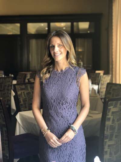Laura Elkman, a caucasian woman with long blondish-brown hair, stands in a Davio's dining room wearing a purple lace dress and colorful bead bracelets