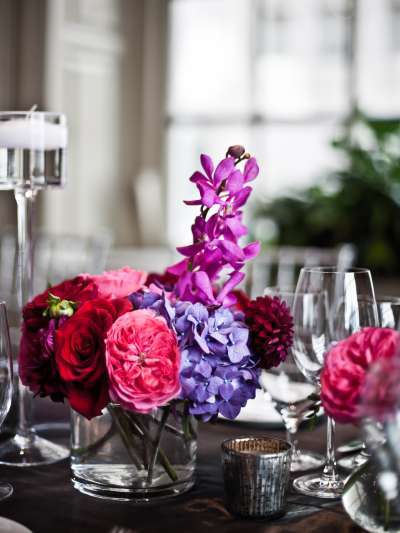 Brightly colored purple, pink, and red flowers are arranged in a glass vase, surrounded by wine glasses and votive candles. A large window is in soft focus in the background.