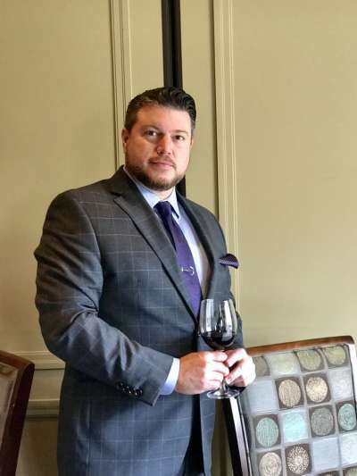 Matthew Rask, a broad-shouldered Caucasian man wearing a gray suit with purple tie and pocked square, stands in the Davio's dining room holding a glass of red wine