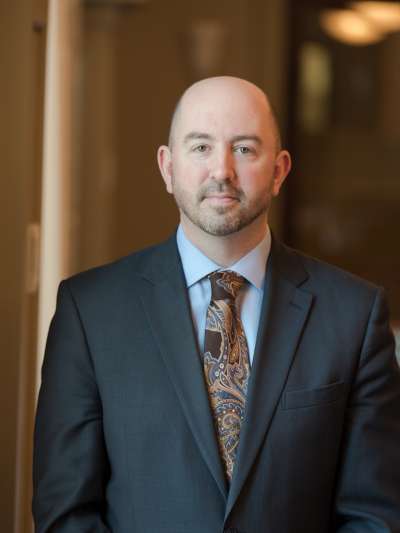 Matthew Del Papa, a bald man with beard stubble, stands in the Davio's dining room wearing a dark gray suit, blue shirt, and paisley tie