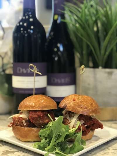 Two kobe meatball sliders with shaved parmesan sit on a square plate with a small arugula salad. Behind them in soft focus are a bottle of Cabernet Sauvignon and a bottle of Pinot Noir, both with purple lables with the Davio's logo, and a green grassy plant in a distressed metallic pot