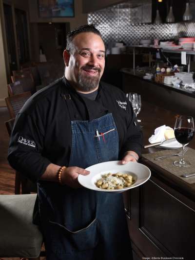 A smiling, heavy-set Caucasian man with a close-cut dark beard, stands in the Davio's Atlanta kitchen wearing a black chef's top and a denim apron, holding a plate of gnocchi topped with parmesan.
