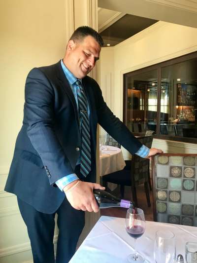 Nick Picozzi, a Caucasian man with close-cropped brown hair, wearing a dark blue suit, blue plaid shirt and striped tie, smiles as he pours red wine in the dining room