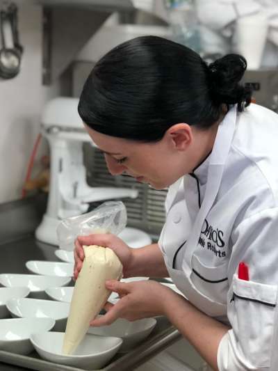 Danielle Roberts, a Caucasian woman with shiny dark hair pulled back into a tight bun, pipes cream into individual dessert bowls