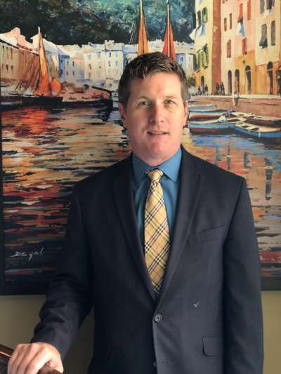 Paul Dunn, in a dark suit with blue shirt and yellow tie, stands in front of a colorful painting of a Mediterranean town on the water