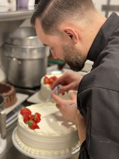 Man dark hair, black jacket, decorating a white cake with strawberries