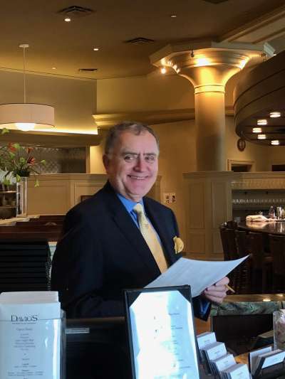 Robert Regan, a smiling middle-aged man wearing a dark suit, blue shirt, and yellow tie, checks on reservations at the host's station