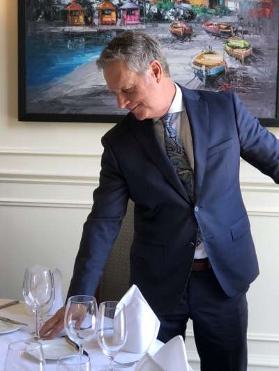 Scott McCully, a Caucasian man with grey hair, wears a blue suit and paisley tie as he leans to adjust a place setting in the Davio's Irvine dining room