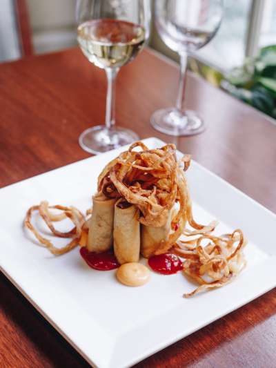 Davio's Philly Cheesesteak Spring Rolls are arranged in a cluster, standing on end on a square, white plate. Skinny onion rings cascade from the top, and dots of spicy ketchup and spicy mayo are around the base. A glass of white wine is in soft focus behind hte plate.