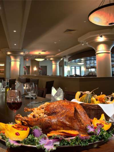 A roast turkey on a platter with winter squash, cranberries, and kale sit on a long table inside Davio's. A glass of wine and basket of gourds are seen behind the platter.