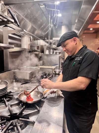Man in kitchen, black jacket, dark hat, pouring sauce into a pan