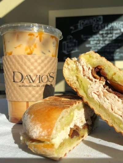 One half of a turkey and bacon panini is propped on the other, sitting next to a latte in a to go cup with a Davio's-labeled cardboard sleeve. A chalkboard menu is in soft focus  in the background