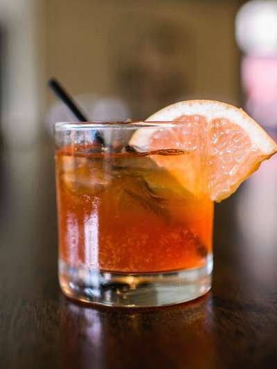 A cocktail in a rocks glass with orange wedge sits on the bar, background in soft focus