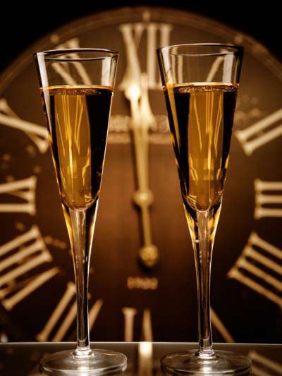 Two flutes of champagne in front of a large clock with roman numerals, hands pointing to one minute before midnight