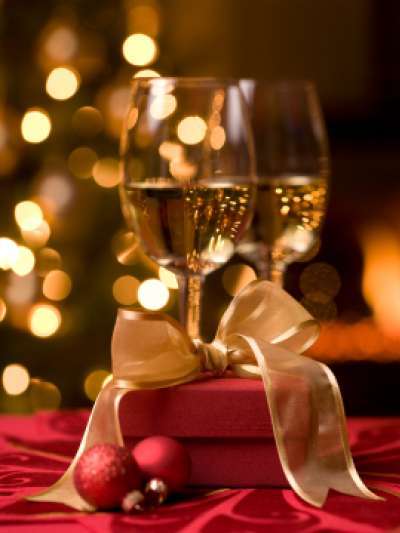 Two glasses of white wine, a red gift box tied with gold ribbon, and two small red ornaments sit on a red tablecloth, with a Christmas tree with white lights and a fire in a fireplace out of focus in the background