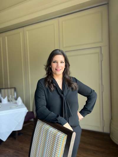 Woman, dark long hair, black shirt, black pants standing behind a chair in dining room