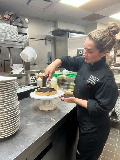 Woman Blond Hair in bun, black chef Coat, decorating a cake