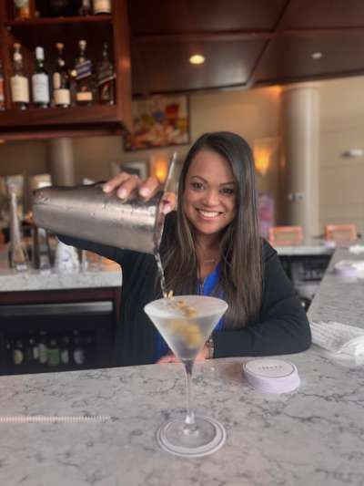 Woman, Dark Hair, Black Jacket, Blue Shirt Pouring a Martini into a Glass