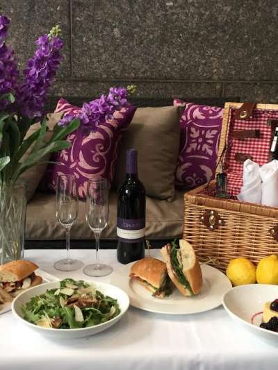 A table with an open picnic basket, two sandwiches, arugula salad, blackberry flan, a bottle of Davio's-labeled wine, and an arrangement of flowers