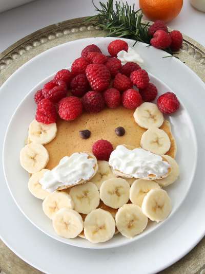 A pancake decorated to look like Santa Claus, with raspberries for his cap, banana slices for his hair and beard, chocolate chips for eyes, a raspberry nose, and a whipped cream mustache