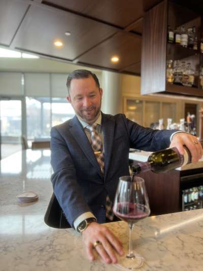 Man, blue suit, short hair, pouring a glass of wine behind the bar