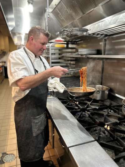 Man in Kitchen, White chef coat, black apron, spooning out pasta with red sauce