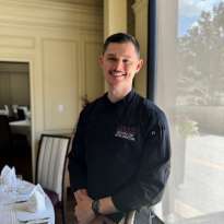 Man, Dark Hair, Mustache, Black Chef Coat standing behind a table