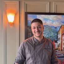 Man, short hair, grey Chef Jacket standing next to a chair 
