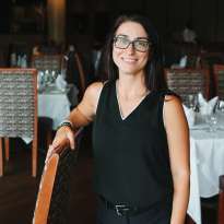 Woman, Black Shirt, Dark Hair, Glasses, Standing with arm leaning on a chair